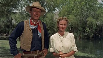 Movie still from “Hondo” (1953), directed by John Farrow – An older man standing next to an older woman in front of trees; Medium shot, Low angle