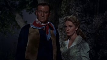 Movie still from “Hondo” (1953), directed by John Farrow – A man and a woman standing next to each other in front of a tree; Medium shot, Low angle