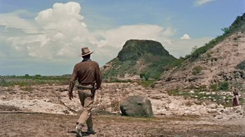 Movie still from “Hondo” (1953), directed by John Farrow – A man in a hat walking on a dirt field; Wide shot, Low angle