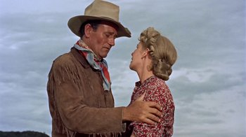 Movie still from “Hondo” (1953), directed by John Farrow – An older man and a younger woman embracing each other in front of a cloudy sky; Medium shot, Low angle