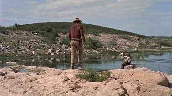 Movie still from “Hondo” (1953), directed by John Farrow – A man standing on top of a hill near a body of water; Extreme Wide shot, Over the shoulder angle
