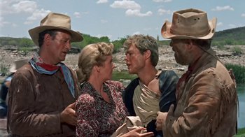 Movie still from “Hondo” (1953), directed by John Farrow – A group of men and a woman talking to each other; Medium shot, Over the shoulder angle