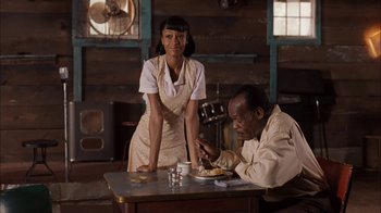 Movie still from “Honeydripper” (2007), directed by John Sayles – A woman standing next to a man at a dinner table; Medium shot, Over the shoulder angle