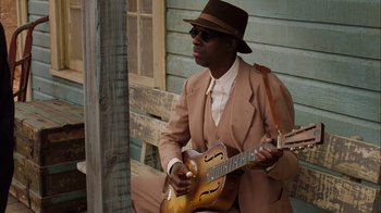 Movie still from “Honeydripper” (2007), directed by John Sayles – A man in a brown suit and hat playing a guitar; Medium shot, High angle