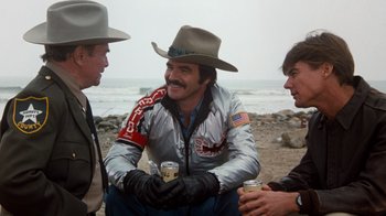 Movie still from “Hooper” (1978), directed by Hal Needham – A man in a cowboy hat sitting next to two other men on the beach; Medium shot, Over the shoulder angle