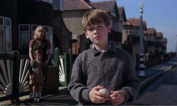 Movie still from “Hope and Glory” (1987), directed by John Boorman – A young boy holding a ball while standing on the street; Medium shot, Low angle