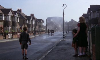 Movie still from “Hope and Glory” (1987), directed by John Boorman – A group of people walking down a street near buildings; Wide shot, Over the shoulder angle