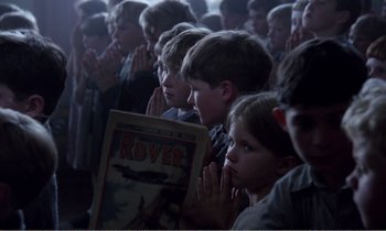Movie still from “Hope and Glory” (1987), directed by John Boorman – A group of people sitting in front of a crowd; Medium shot, High angle