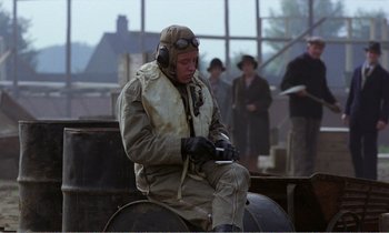 Movie still from “Hope and Glory” (1987), directed by John Boorman – A man in a flight suit sitting on top of a barrel; Wide shot, High angle
