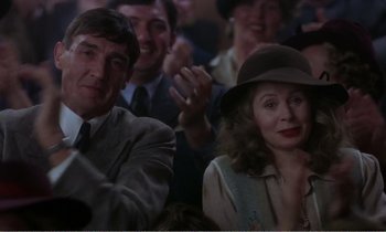 Movie still from “Hope and Glory” (1987), directed by John Boorman – A group of people sitting next to each other in a room; Medium shot, Over the shoulder angle