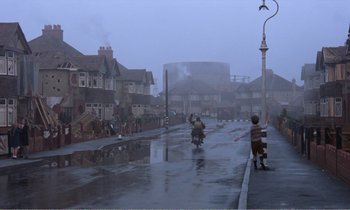 Movie still from “Hope and Glory” (1987), directed by John Boorman – Two people riding motorcycles down a street in the pouring rain; Extreme Wide shot, Over the shoulder angle