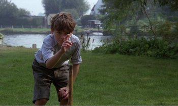 Movie still from “Hope and Glory” (1987), directed by John Boorman – A young boy holding a baseball bat on a field; Medium shot, Over the shoulder angle