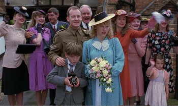 Movie still from “Hope and Glory” (1987), directed by John Boorman – A group of people standing next to each other holding flowers; Medium shot, Over the shoulder angle