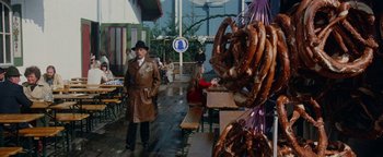 Movie still from “Hopscotch” (1980), directed by Ronald Neame – A man in a brown raincoat standing in front of some pretzels; Wide shot, High angle