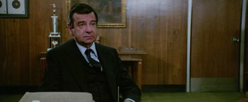 Movie still from “Hopscotch” (1980), directed by Ronald Neame – An older man wearing a suit and tie sitting at a table; Medium shot, Over the shoulder angle
