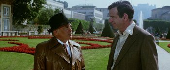 Movie still from “Hopscotch” (1980), directed by Ronald Neame – A man in a brown jacket and a man in a brown hat talking to each other in a garden; Medium shot, High angle