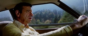 Movie still from “Hopscotch” (1980), directed by Ronald Neame – A man driving a car on a road with trees in the background; Close Up shot, Low angle