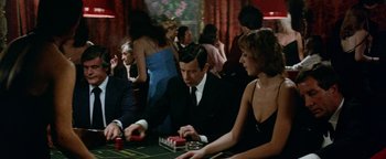 Movie still from “Hopscotch” (1980), directed by Ronald Neame – A group of people sitting at a table playing a game; Medium shot, Over the shoulder angle