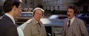 Movie still from “Hopscotch” (1980), directed by Ronald Neame – A man in a trench coat standing in front of a car; Medium shot, Over the shoulder angle