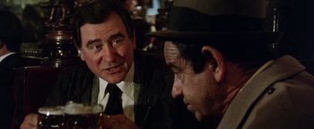 Movie still from “Hopscotch” (1980), directed by Ronald Neame – Two men are sitting at a table and one of them is holding a drink; Close Up shot, Over the shoulder angle