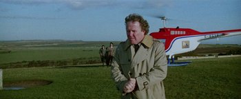 Movie still from “Hopscotch” (1980), directed by Ronald Neame – A man in a trench coat standing next to a helicopter; Medium shot, Over the shoulder angle