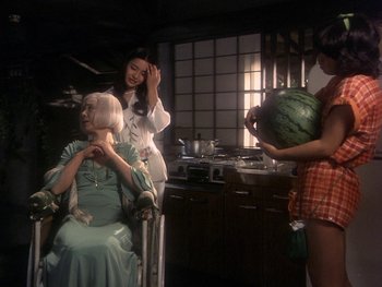 Movie still from “House” (1977), directed by Nobuhiko Ôbayashi – A woman sitting in front of a woman holding a watermelon in a kitchen; Wide shot, High angle