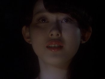 Movie still from “House” (1977), directed by Nobuhiko Ôbayashi – A young woman with a dark background looking at the camera; Close Up shot, Over the shoulder angle