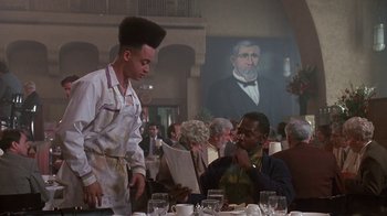 Movie still from “House Party 2” (1991), directed by Doug McHenry – A man standing next to a man sitting at a dining table; Medium shot, Low angle