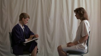 Movie still from “House of Games” (1987), directed by David Mamet – Two people are sitting in a chair and one of them is writing; Medium shot, Over the shoulder angle