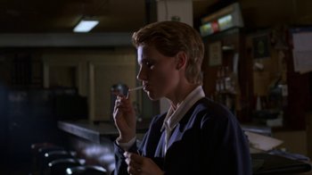 Movie still from “House of Games” (1987), directed by David Mamet – A man holding a cigarette in his mouth while standing; Medium shot, Low angle