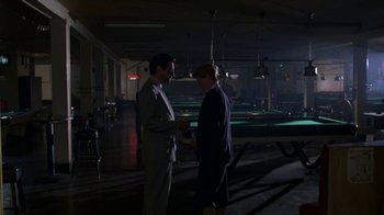 Movie still from “House of Games” (1987), directed by David Mamet – Two men standing in a dark room talking to each other; Wide shot, Over the shoulder angle