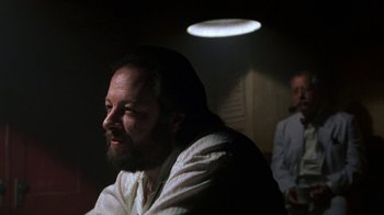 Movie still from “House of Games” (1987), directed by David Mamet – A man with a long beard is sitting in a dark room; Close Up shot, Low angle