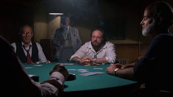 Movie still from “House of Games” (1987), directed by David Mamet – A man sitting at a table in front of a group of people; Medium shot, Over the shoulder angle