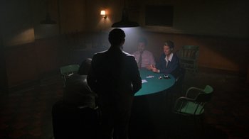 Movie still from “House of Games” (1987), directed by David Mamet – A group of people sitting at a table in a dark room; Wide shot, Over the shoulder angle
