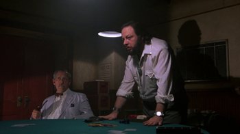 Movie still from “House of Games” (1987), directed by David Mamet – A man standing at a table with another man sitting at a table; Medium shot, Over the shoulder angle
