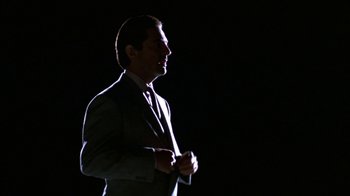 Movie still from “House of Games” (1987), directed by David Mamet – A man in a suit and tie standing in front of a black background; Medium shot, Low angle