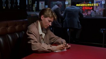 Movie still from “House of Games” (1987), directed by David Mamet – A man sitting at a table writing on a piece of paper; Medium shot, High angle