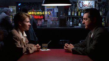 Movie still from “House of Games” (1987), directed by David Mamet – Two people sitting at a table in front of a glass of whiskey; Medium shot, Over the shoulder angle
