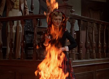 Movie still from “House of Wax” (1953), directed by André De Toth – A woman standing in front of a fire on top of a staircase; Medium shot, Over the shoulder angle