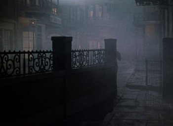 Movie still from “House of Wax” (1953), directed by André De Toth – A person standing on a sidewalk in the fog; Wide shot, High angle