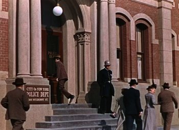 Movie still from “House of Wax” (1953), directed by André De Toth – A man in a police uniform standing in front of a building; Wide shot, Low angle