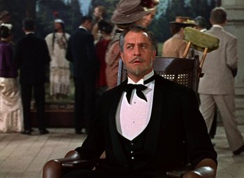 Movie still from “House of Wax” (1953), directed by André De Toth – A man in a suit sitting in a chair in front of a crowd of onlookers; Medium shot, Low angle