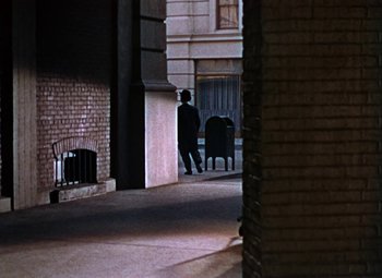 Movie still from “House of Wax” (1953), directed by André De Toth – A man standing in a room next to a brick wall; Extreme Wide shot, High angle