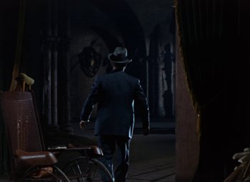 Movie still from “House of Wax” (1953), directed by André De Toth – A man in a suit and hat walking down a dark hallway; Wide shot, Over the shoulder angle