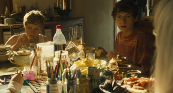 Movie still from “How I Live Now” (2013), directed by Kevin Macdonald – A young man sitting at a table with food and drinks on it; Medium shot, High angle