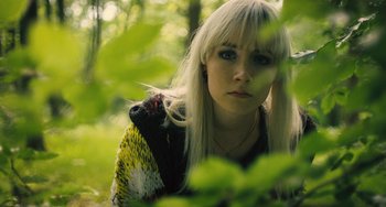 Movie still from “How I Live Now” (2013), directed by Kevin Macdonald – A woman with blonde hair and a scarf around her neck looks at the camera; Close Up shot, High angle