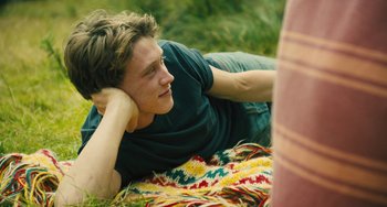 Movie still from “How I Live Now” (2013), directed by Kevin Macdonald – A man laying in the grass with his hands behind his head; Medium shot, Over the shoulder angle