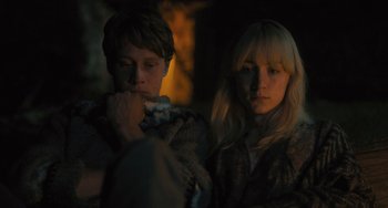 Movie still from “How I Live Now” (2013), directed by Kevin Macdonald – A man and a woman sitting next to each other in front of a fire; Medium shot, Over the shoulder angle