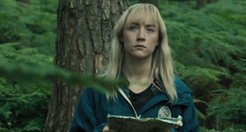 Movie still from “How I Live Now” (2013), directed by Kevin Macdonald – A woman with blonde hair is standing in front of a tree holding a book; Close Up shot, Over the shoulder angle