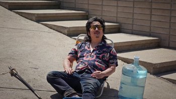 Movie still from “How It Ends” (2021), directed by Daryl Wein – A man sitting on the ground wearing a shirt and sunglasses; Medium shot, High angle